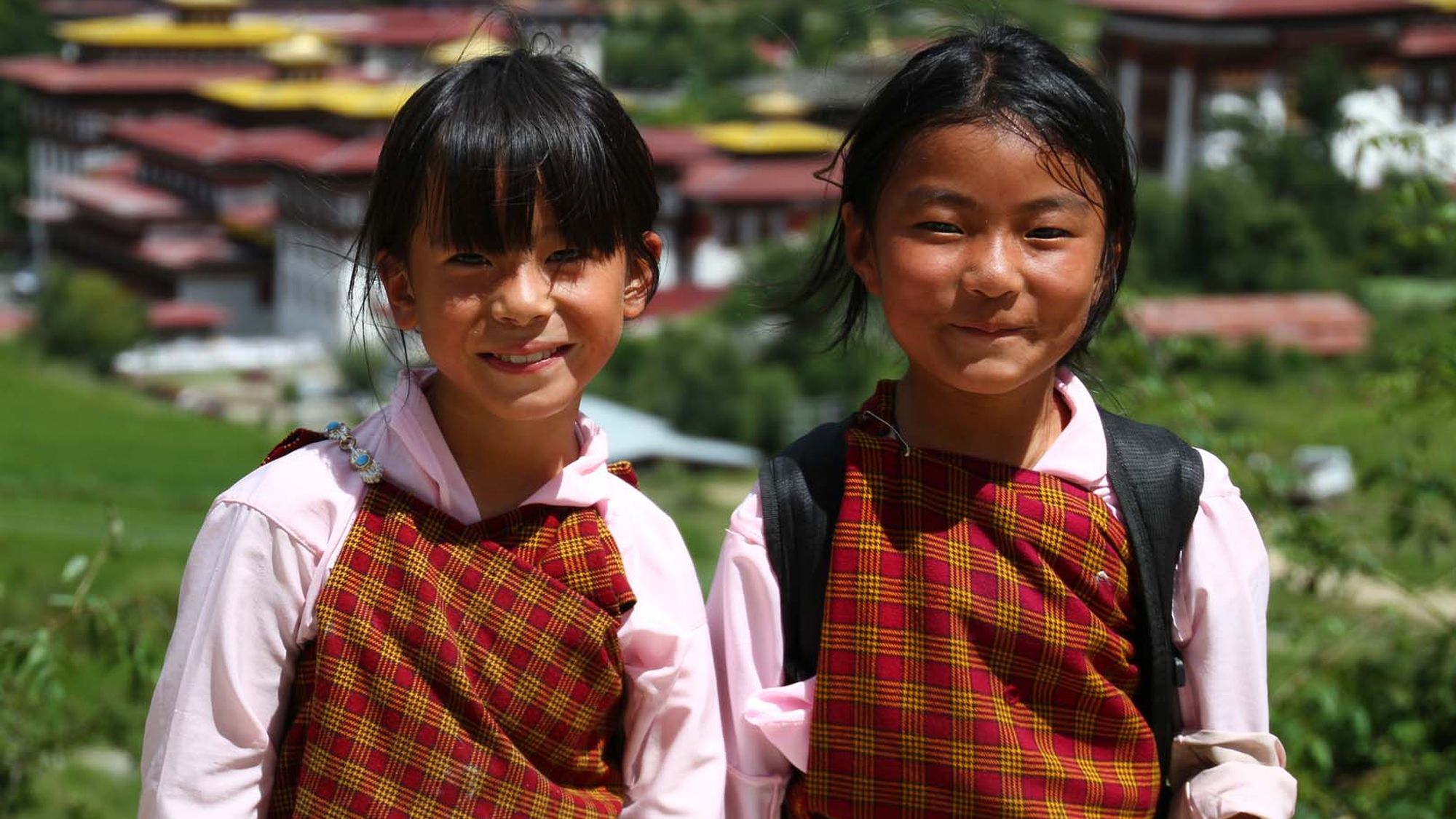 Two Bhutanese schoolgirls in kira standing above a valley of traditional rooftops
